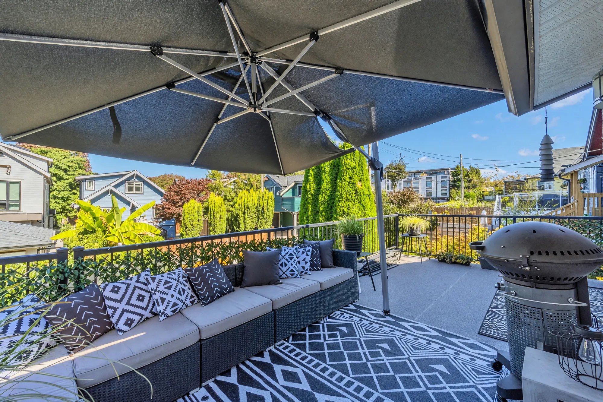 On the balcony under a large umbrella: an outdoor sectional with patterned cushions, an outdoor rug, a barbecue, the railing and the rooftops beyond