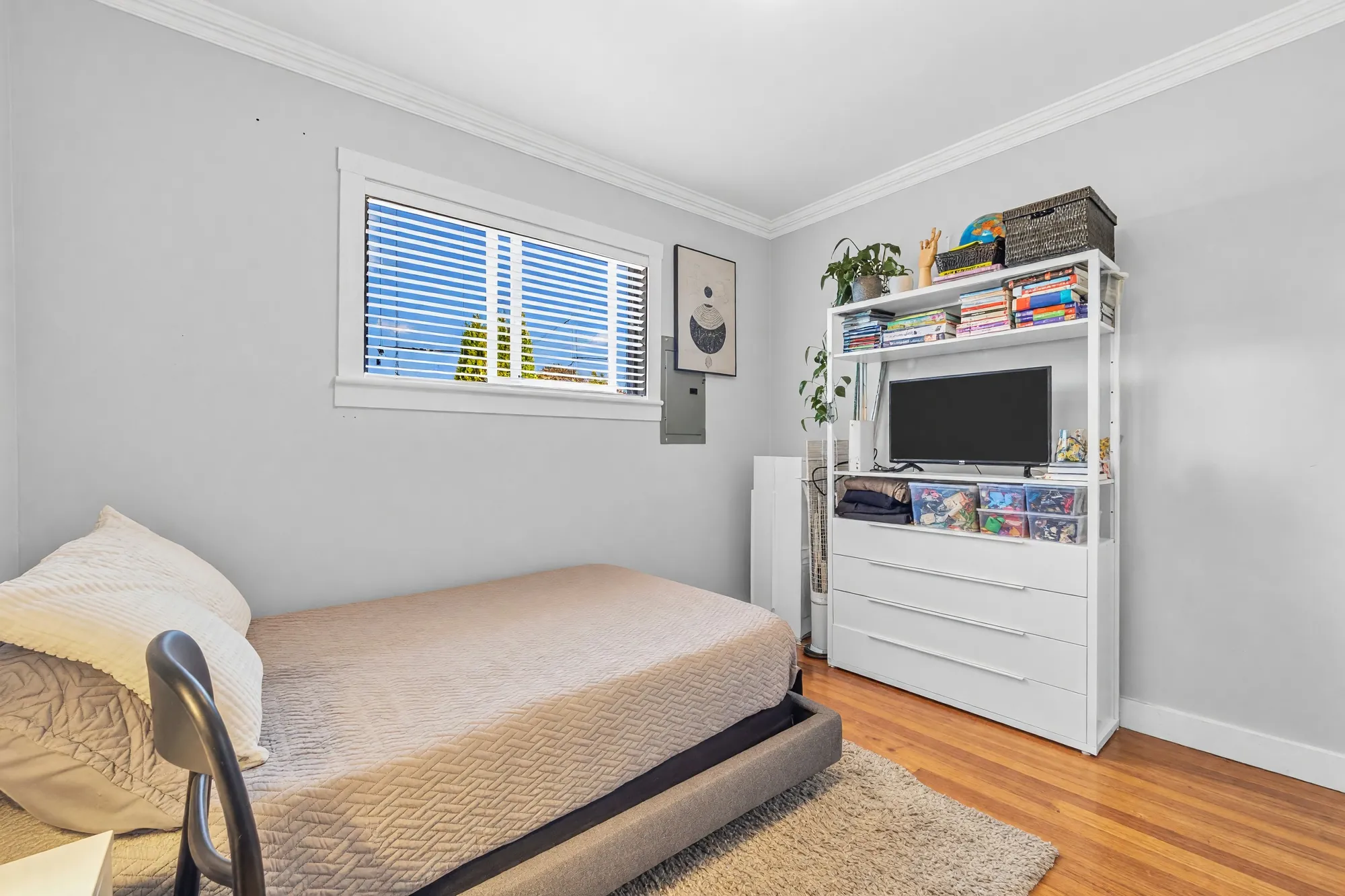 The second bedroom: a bed, a white shelving unit with a television, a window with blinds, hardwood