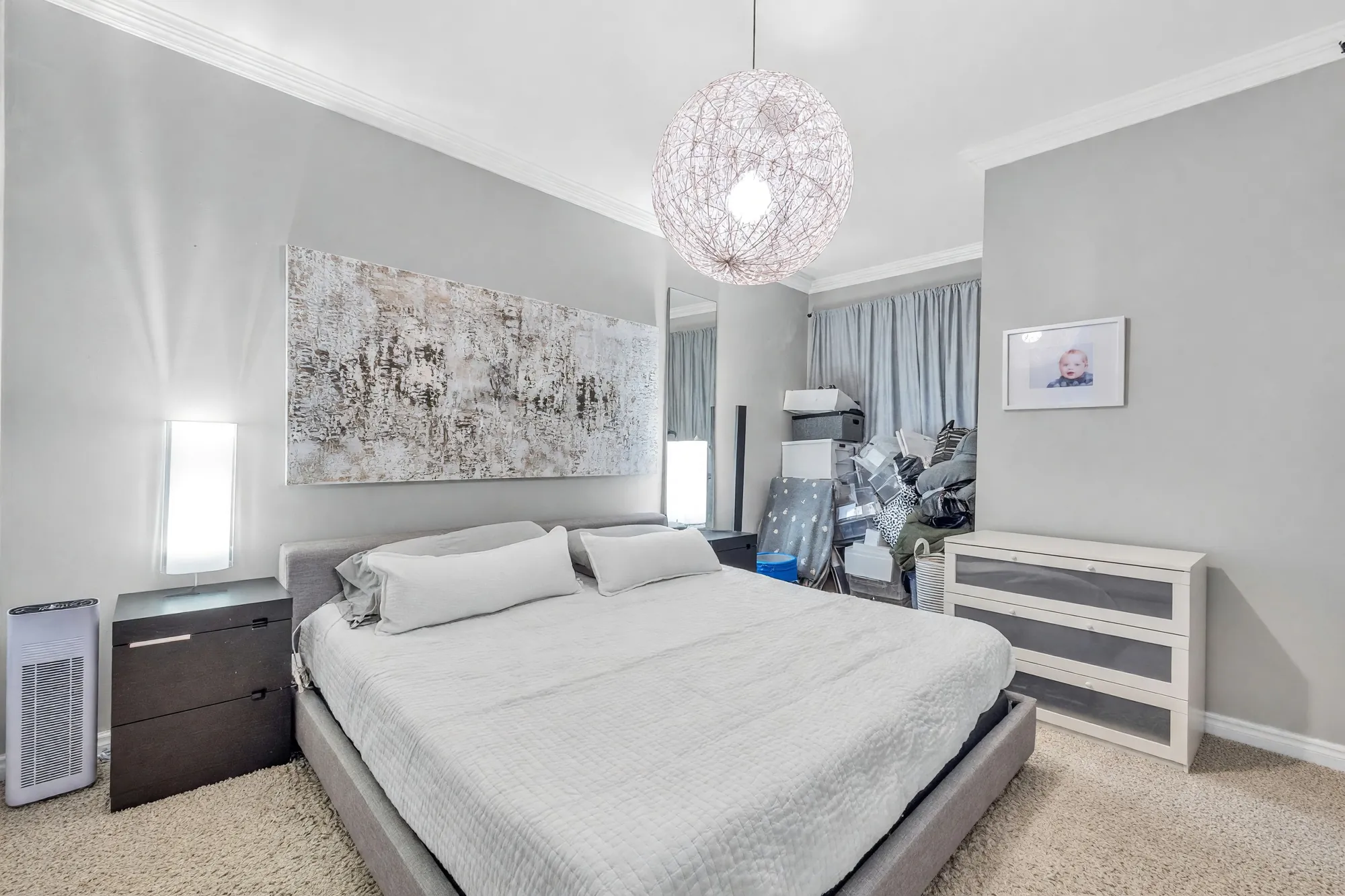 The larger bedroom: a bed under a wire-globe pendant, a large abstract canvas, curtains, a white dresser, carpet