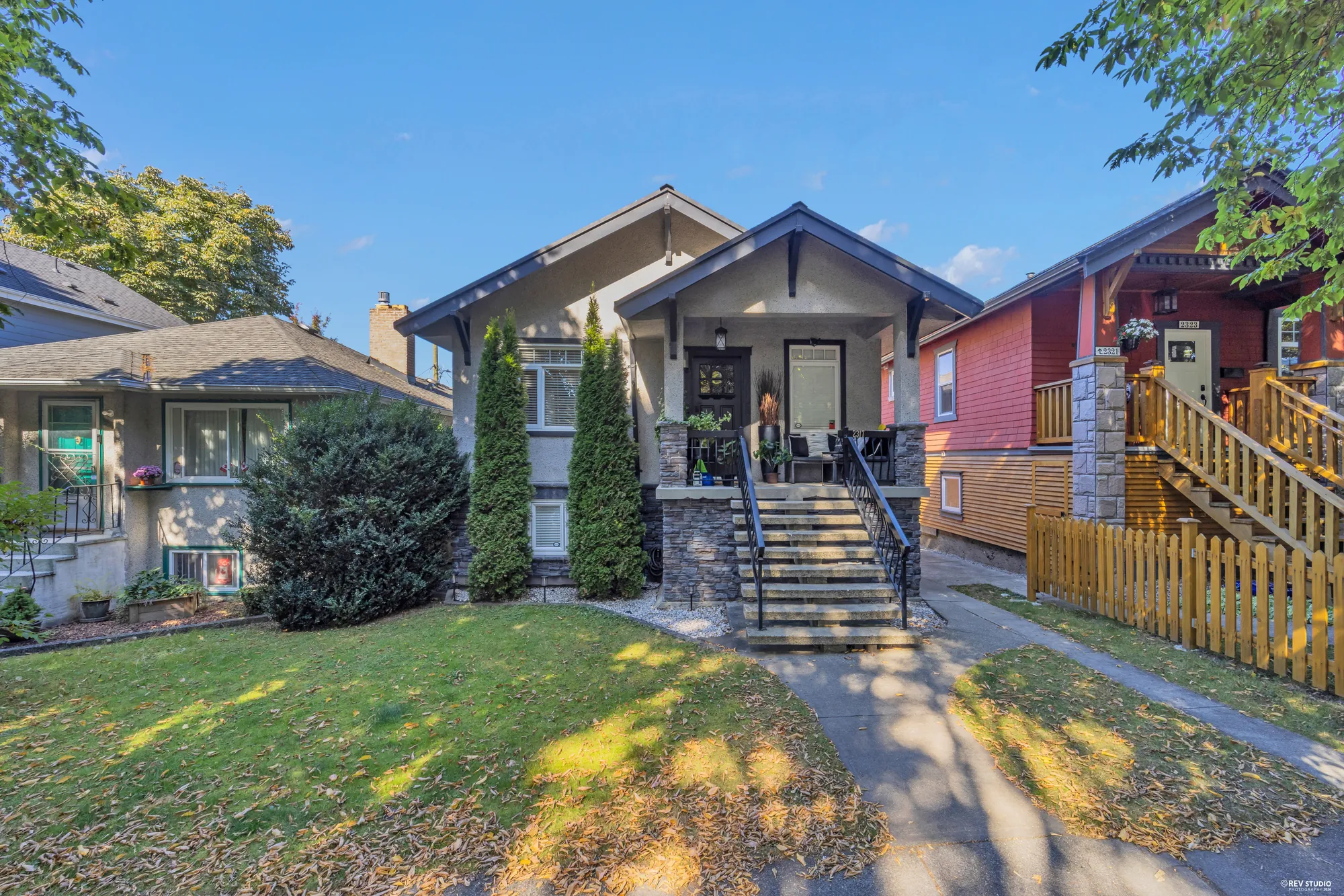 The front of the house as photographed: grey stucco, a gabled porch on stone piers, two columnar cedars, the lawn brown and covered in fallen leaves, the red house next door