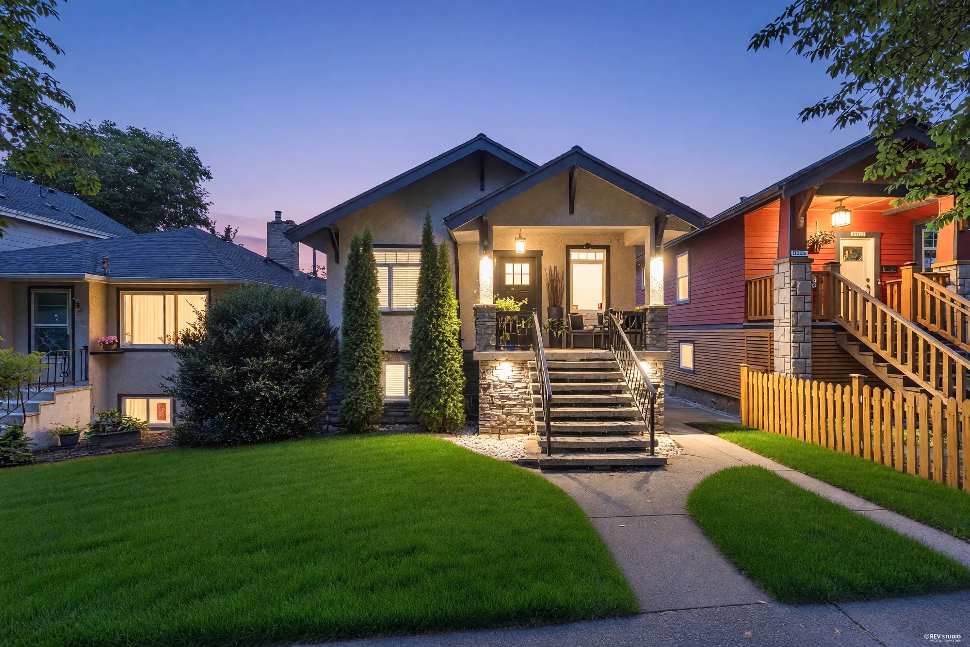 2317 Graveley Street at dusk: a grey stucco Craftsman with a gabled porch on stone piers, two columnar cedars either side of the steps, the porch and windows lit