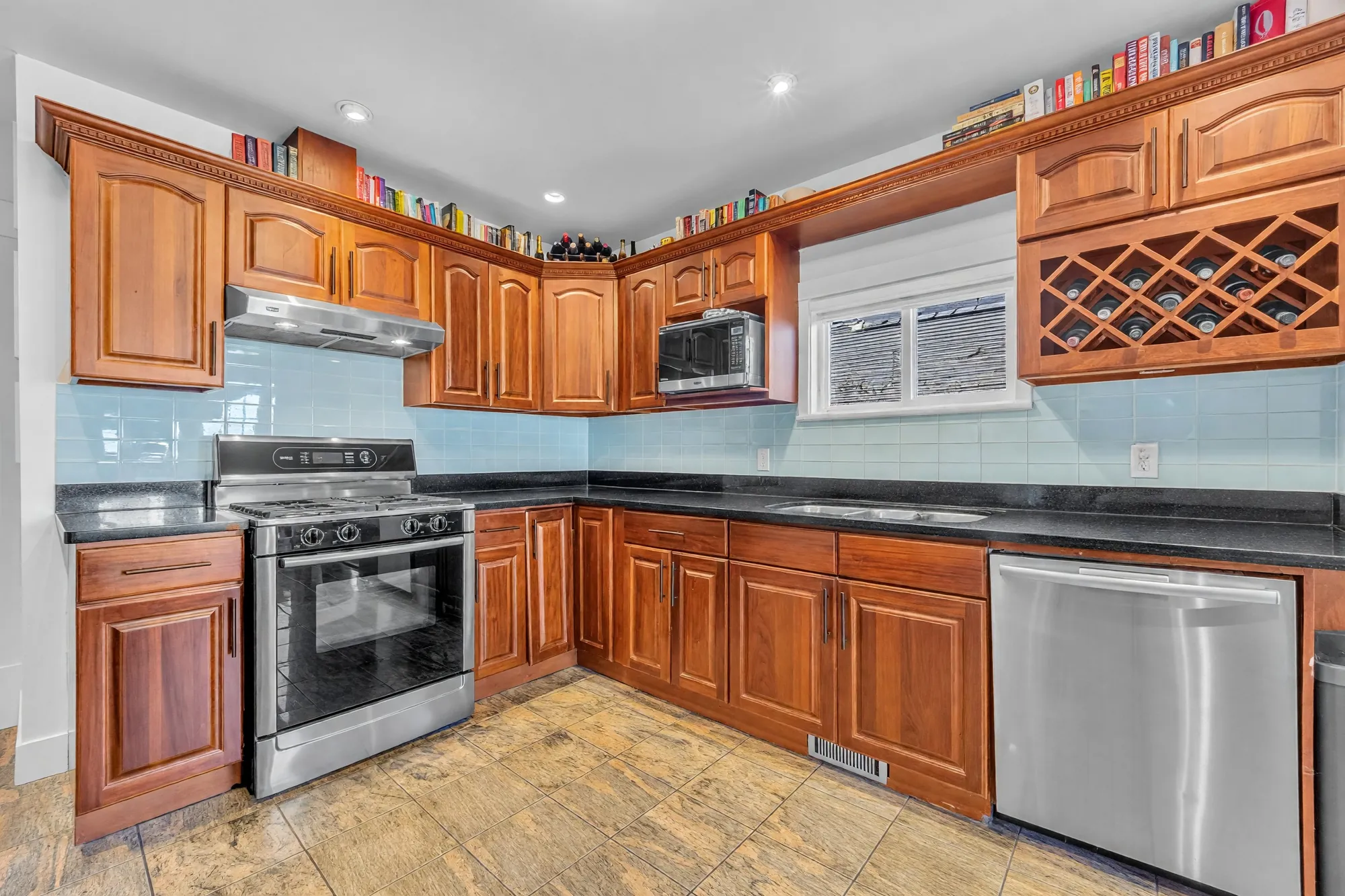 The kitchen: cherry cabinets, dark stone counters, a pale blue glass-tile backsplash, a stainless range and dishwasher, a wine rack over the window