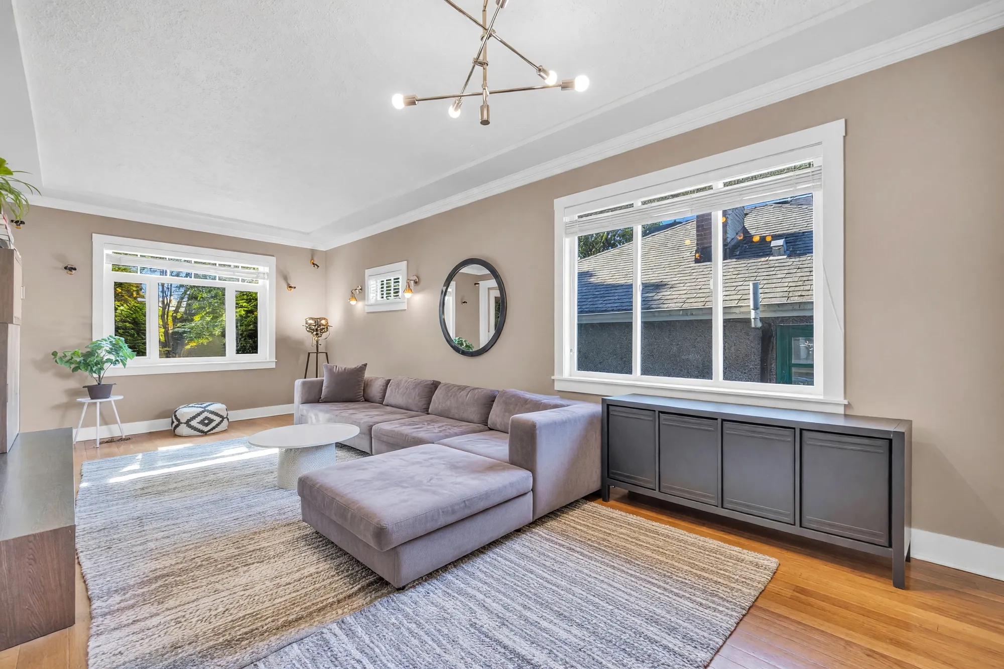 The living room toward the front windows: a grey sectional, an oval mirror, black cabinets under the side window, a chandelier, hardwood