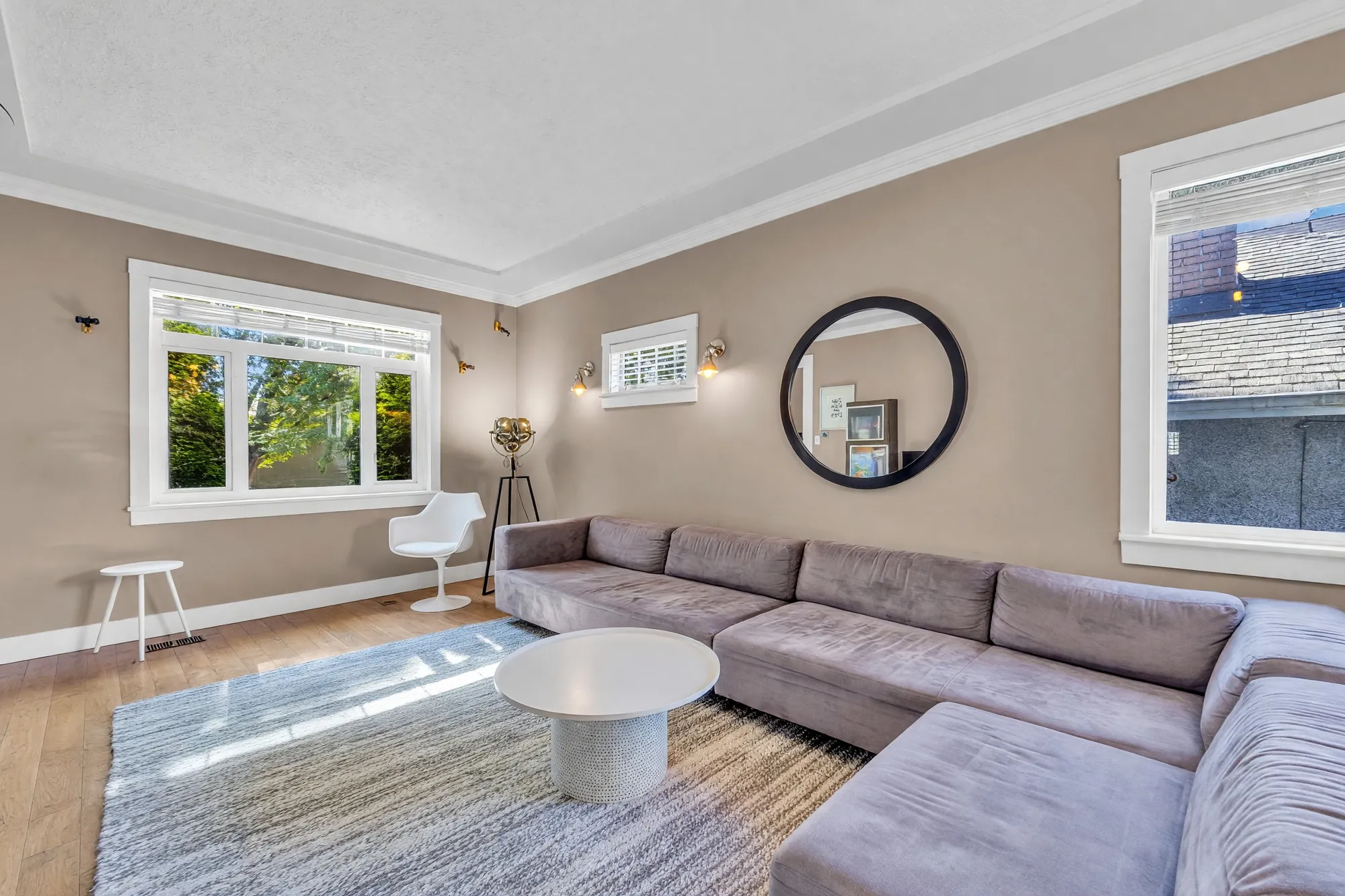 The sectional under the side windows, a white chair by the window, the oval mirror