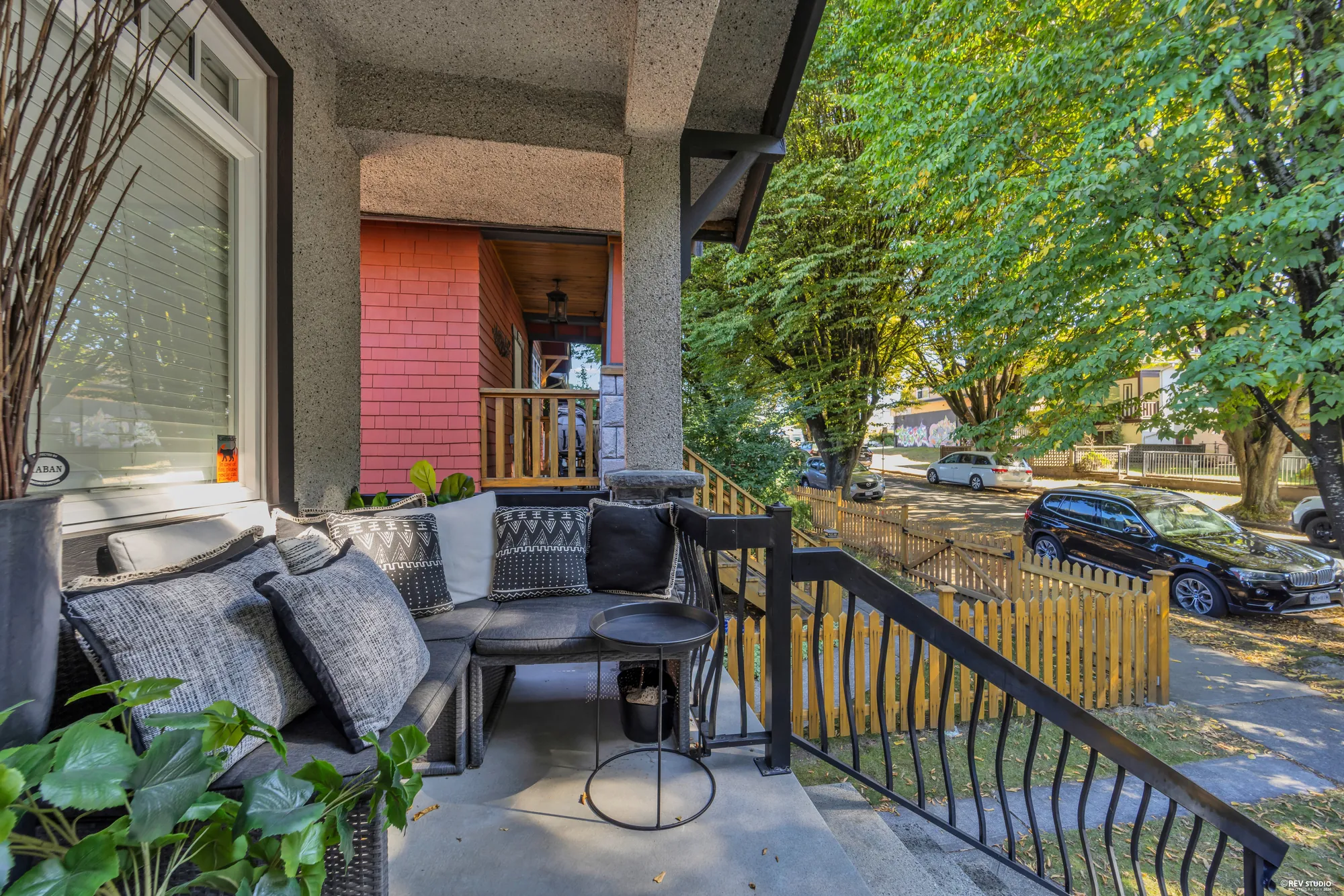 On the porch: an outdoor sofa with cushions, the railing, the street trees and the houses opposite